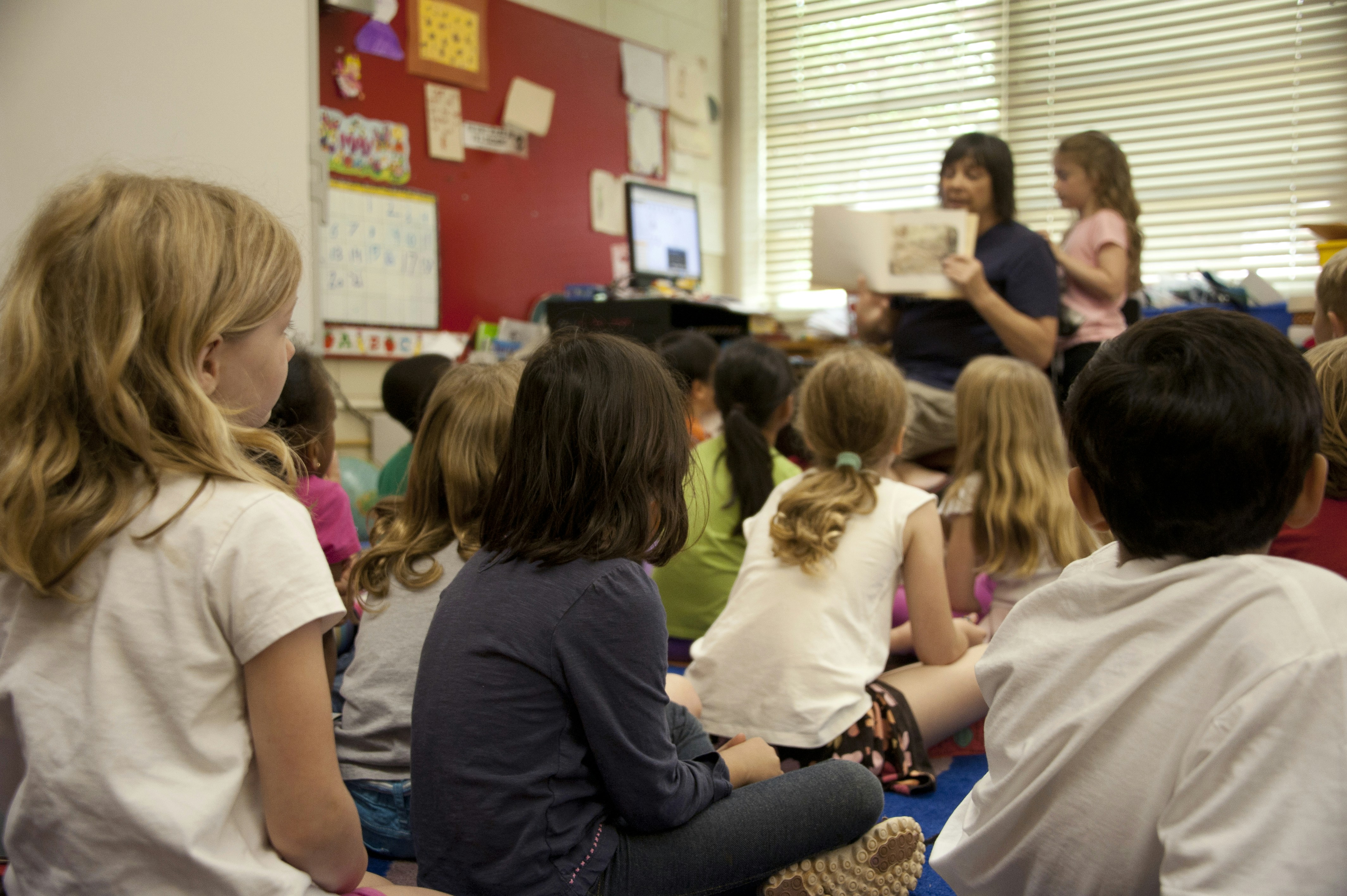 Captured in a metropolitan Atlanta, Georgia primary school, this photograph depicts a typical classroom scene, where an audience of school children were seated on the floor before a teacher at the front of the room, who was reading an illustrated storybook, during one of the scheduled classroom sessions. One of the female students was assisting the teacher, while the rest of the class listened attentively to the instructor’s narrative.  by Teacher Strategies people sitting on chair inside room
